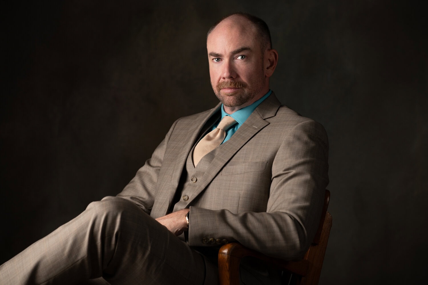 A bald man with a trimmed beard wearing a tan suit, teal shirt, and beige tie sits confidently in a wooden chair, gazing at the camera against a dark, muted background.