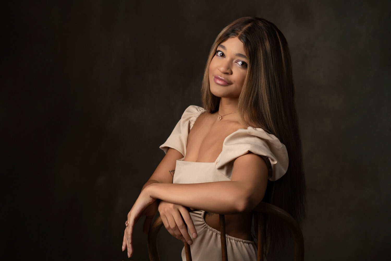 A young woman with long straight hair sits sideways on a wooden chair, smiling gently. She wears a beige dress with puffed sleeves against a dark, neutral background.