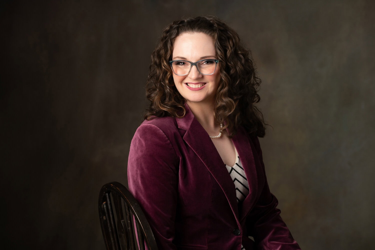 A woman with curly brown hair, wearing glasses, a maroon velvet blazer, a black-and-white striped blouse, and a pearl necklace, sits smiling on a chair against a dark, neutral studio background.