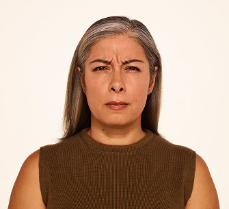 A woman with long gray hair and a brown sleeveless top is looking directly at the camera with a serious, slightly furrowed brow expression.