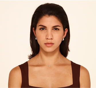 A woman with straight dark hair, wearing a brown sleeveless top, small hoop earrings, and a delicate necklace, stands against a plain light background, looking directly at the camera with a neutral expression.